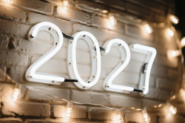 Witte neoncijfers 2027 hangen op een witgeschilderde bakstenen muur, met warme lichtslingers op de voorgrond en achtergrond.