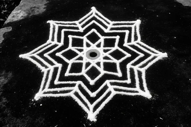 Een zwart-witfoto van bovenaf toont een symmetrisch geometrisch rangolipatroon van wit poeder op een donker stenen oppervlak met structuur.
