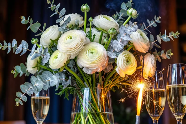 Een glazen vaas met witte ranonkels en zilverkleurig bespoten eucalyptus staat naast brandende sterretjes en champagnecoupes tegen een donkere achtergrond.