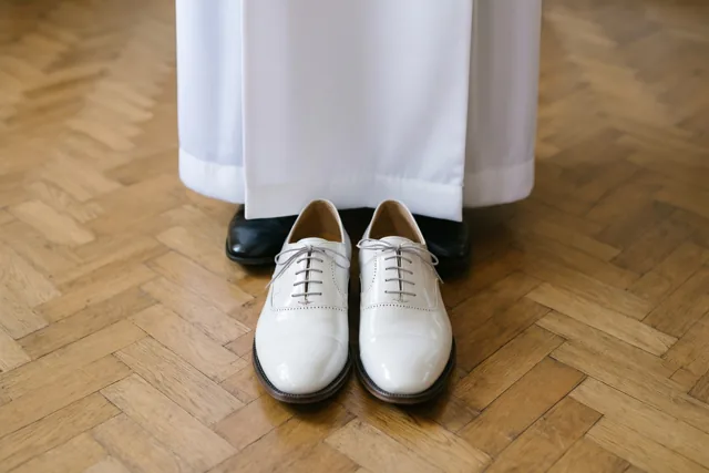 Een paar witte lakschoenen staat op een houten parketvloer naast een wit communiegewaad.