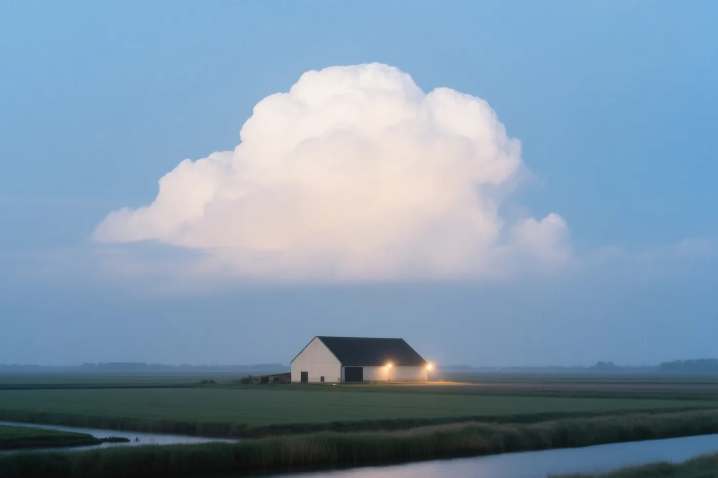 Een witte schuur met een donker dak staat in een vlakke groene polder onder een grote witte wolk aan een blauwe lucht, met een waterloop en riet op de voorgrond.