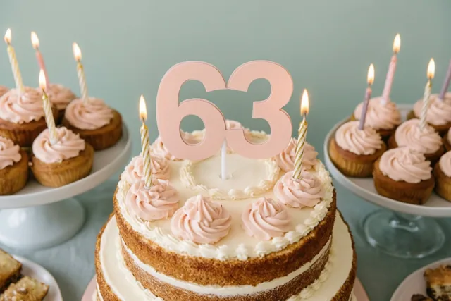 Een taart met meerdere verdiepingen, wit glazuur, roze krullen, een grote roze versiering met 63 en brandende kaarsen staat tussen cupcakes op standaards tegen een blauwgroene achtergrond.