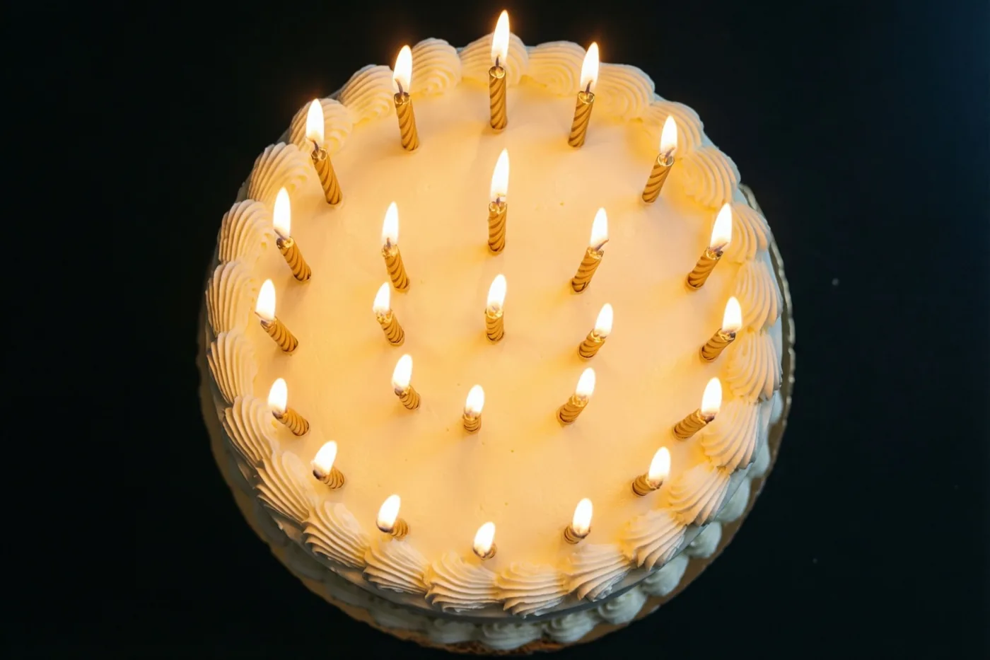 Van bovenaf is een ronde taart met wit glazuur en een kring brandende gouden kaarsen te zien tegen een zwarte achtergrond.
