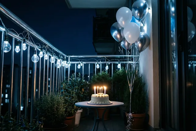 Een kleine witte taart met brandende kaarsjes staat 's nachts op een ronde tafel op een balkon, tussen lichtslingers en zilveren en witte ballonnen.