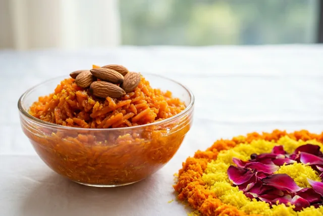 Een glazen kom oranje wortelhalwa met amandelen staat op een witte ondergrond naast een rangoli van oranje en geel poeder en paarse rozenblaadjes, met een raam op de achtergrond.