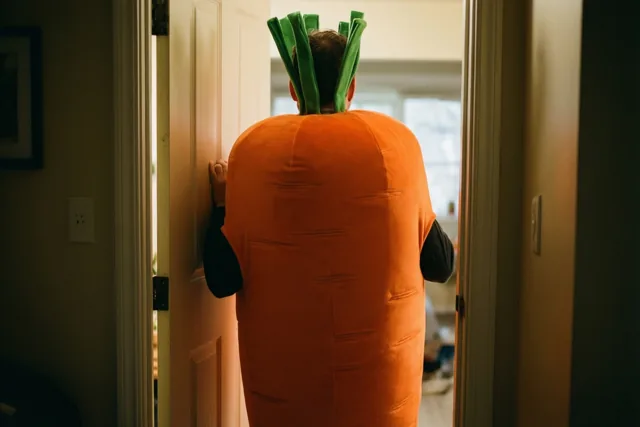 Een man in een groot oranje wortelkostuum met groen loof duwt zich met zijn rug naar de camera door een smalle deuropening in warm licht.