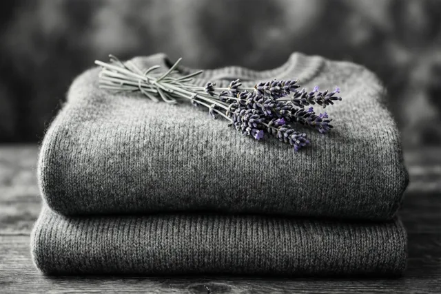Twee opgevouwen kasjmieren truien liggen op elkaar op een houten ondergrond, met een takje lavendel erbovenop in gedempte tinten.