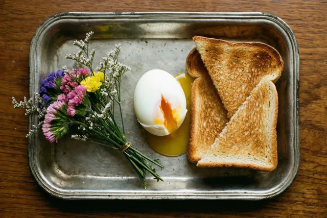 Een oud metalen dienblad op een houten ondergrond bevat een zachtgekookt ei met vloeibare dooier, twee reepjes geroosterd brood en een klein boeket veldbloemen.