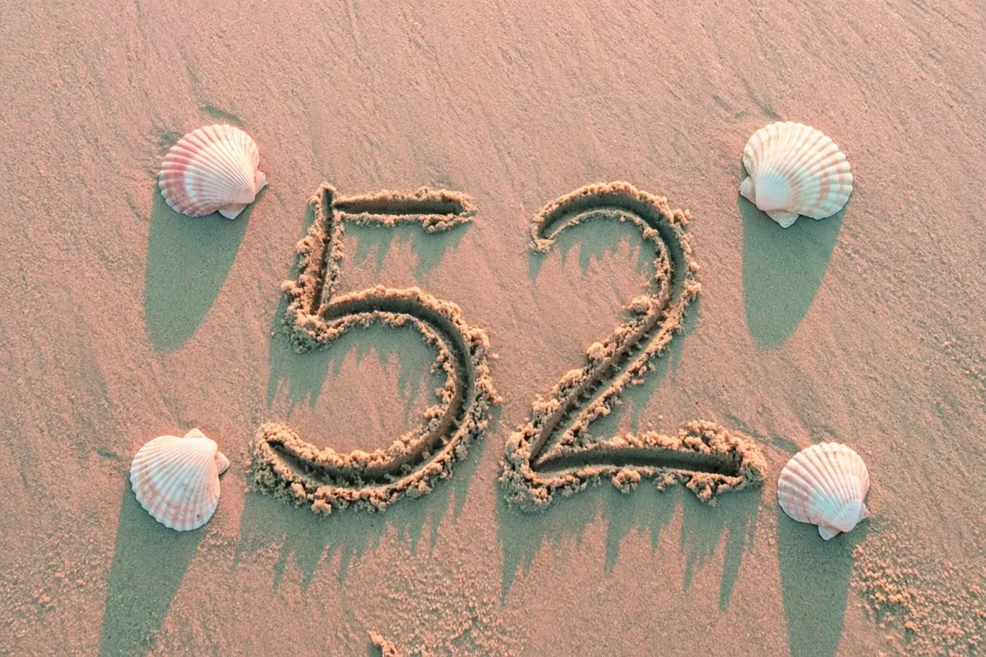 Het getal 52 is in het strandzand getekend tussen schelpen in het roze en oranje licht van de ondergaande zon.