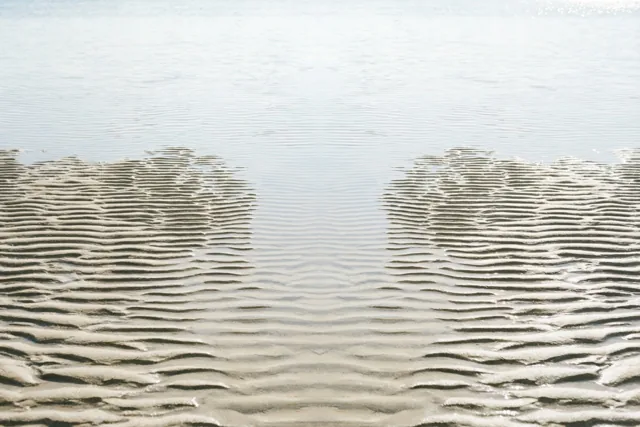Ribbels in beige zand liggen bij eb langs de waterkant, met kalm lichtblauw water op de achtergrond.