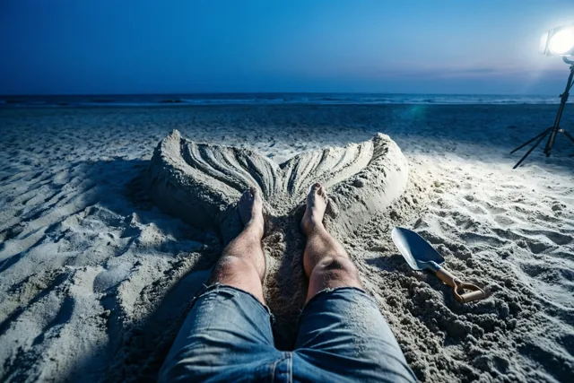 Iemands benen liggen op een strand in de schemering onder zand in de vorm van een zeemeerminnenstaart, verlicht door een felle spot met een schopje ernaast.