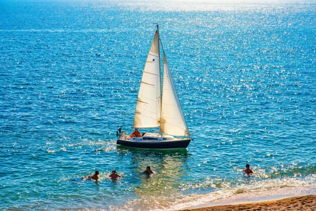 Een kleine zeilboot met witte zeilen vaart over een glinsterende blauwe zee bij een zandstrand, met zwemmers in het ondiepe water langs de oever.