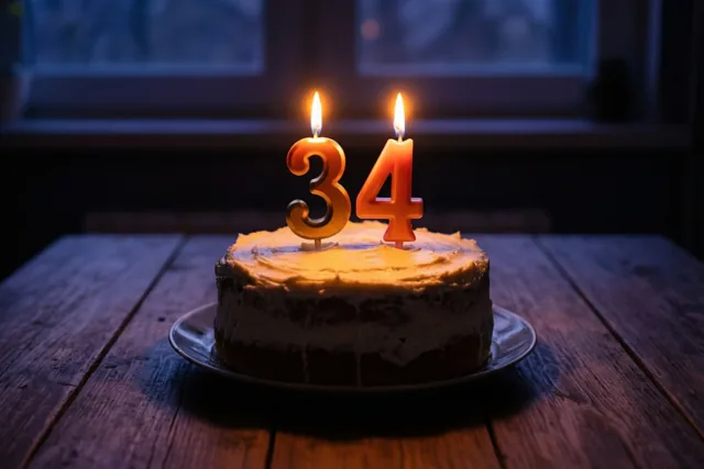 Een zelfgemaakte roomtaart met brandende cijferkaarsen die 34 vormen staat op een houten tafel in warm kaarslicht.