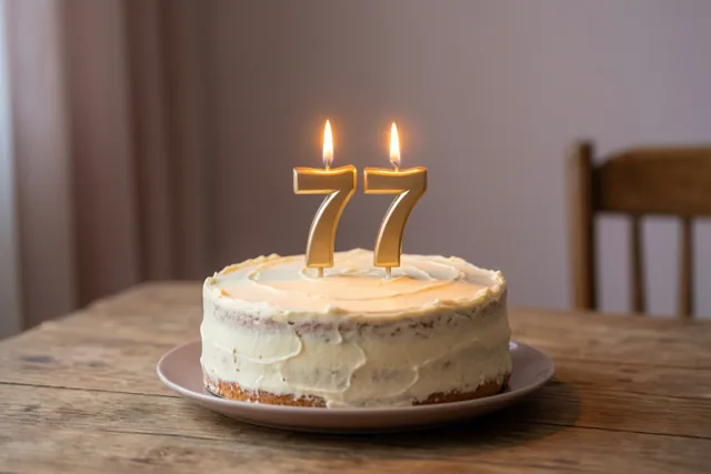 Een zelfgemaakte roomtaart met twee brandende gouden cijferkaarsen die 77 vormen, staat op een houten tafel in warm kaarslicht.