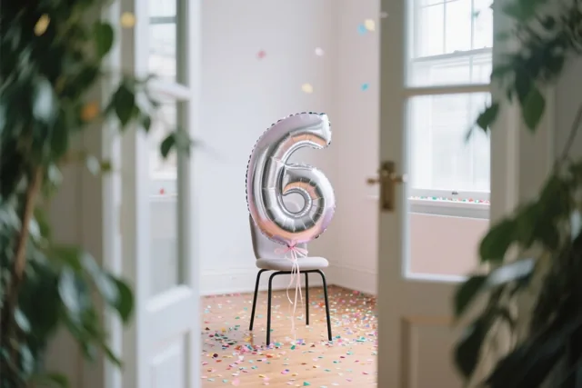 Een zilver- en pastelkleurige ballon in de vorm van het cijfer 6 is aan een stoel vastgemaakt in een lichte kamer met confetti op de houten vloer en groen aan weerszijden.