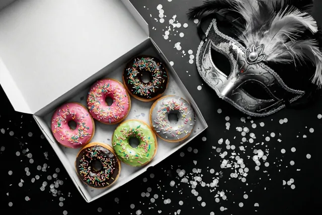 Een open witte doos met zes kleurrijk geglazuurde donuts met strooisel staat naast een zilveren paillettenmasker met veren op een zwarte ondergrond met witte confetti.