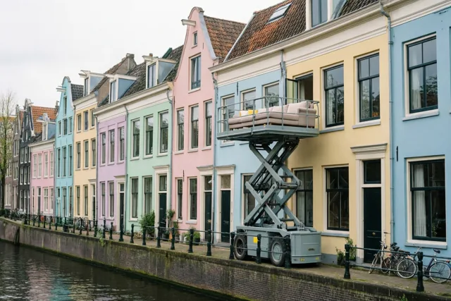 Een grijze schaarlift tilt een beige zetel naar een balkonraam bij pastelkleurige huizen die weerspiegelen in een kalm kanaal onder een bewolkte hemel.