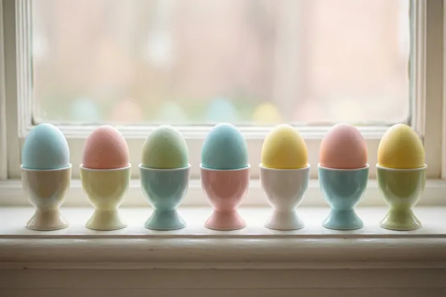 Zeven handbeschilderde eieren in pasteltinten staan in verschillende keramieken eierdopjes op een witte vensterbank in zacht daglicht.