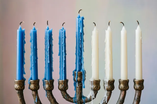 Een sierlijke metalen kandelaar draagt vier blauwe en drie witte smeltende kaarsen, met wasdruppels langs de kaarsen en de armen tegen een roze-blauwe achtergrond.