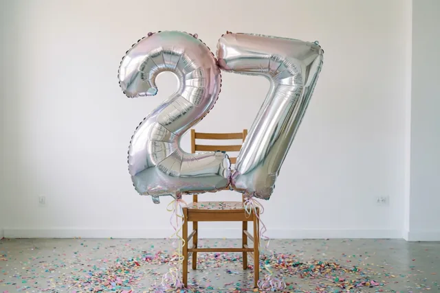 Grote zilver- en pastelkleurige cijferballonnen vormen 27 aan een houten stoel in een lichte sobere kamer, met gekleurde confetti op de vloer.