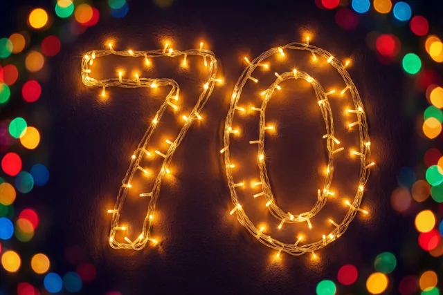 Warme lichtjes vormen het getal 70 op een donkere muur met structuur, omringd door wazige gekleurde feestlichtjes.