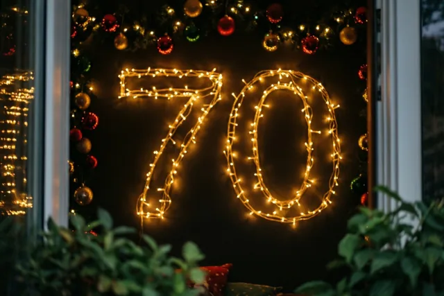 Warme lichtjes vormen 70 op een donkere muur, omlijst door een raam met wazige feestlichtjes en kerstversiering.