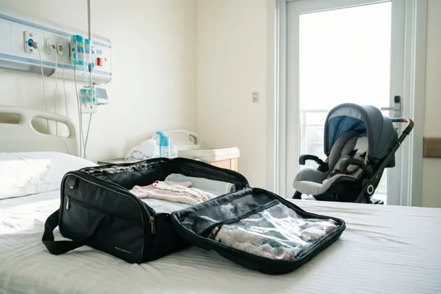 Een open zwarte ziekenhuistas met babykleertjes staat op een ziekenhuisbed, met een autostoeltje voor een baby bij de deur en medische apparatuur in de lichte witte kamer.