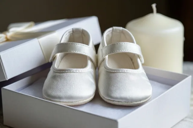 Een paar witte zijden babyschoentjes ligt in een witte geschenkdoos naast een witte doopkaars in zacht licht tegen een neutrale achtergrond.