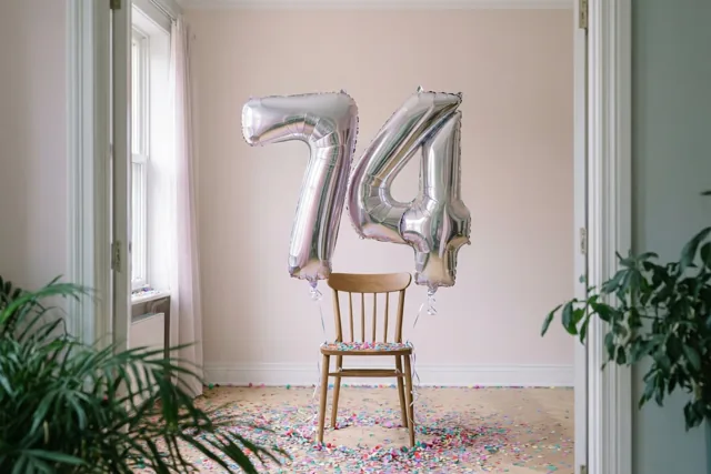 Zilveren folieballonnen vormen 74 aan een houten stoel in een lichte kamer met pastelkleurige muren, kleurrijke confetti en groene planten.