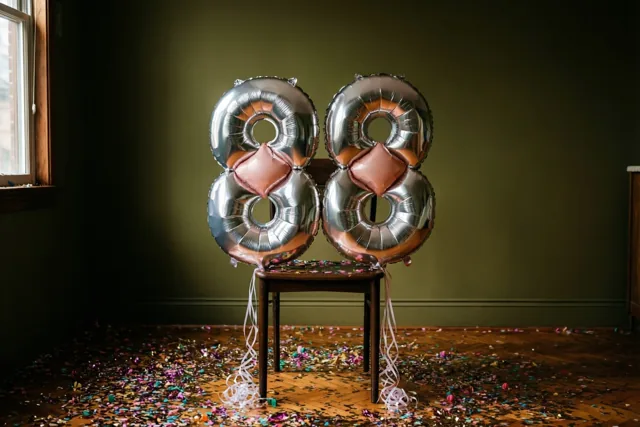 Twee zilveren folieballonnen in de vorm van een 8 zijn vastgemaakt aan een houten stoel in een donkergroene kamer met confetti op de vloer en daglicht van links.