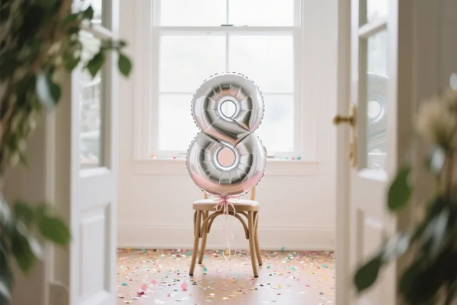 Een zilveren ballon met pasteltinten in de vorm van een 8 is vastgemaakt aan een houten stoel in een lichte kamer met confetti op de vloer, gezien door open deuren met groen aan weerszijden.