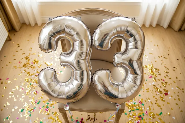Twee grote zilverkleurige folieballonnen vormen 33 aan een beige stoel in een lichte kamer, met gouden en gekleurde confetti op de houten vloer.