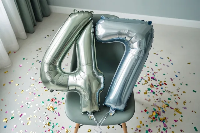 Twee grote zilveren folieballonnen in de vorm van 4 en 7 zijn vastgebonden aan een grijze stoel in een lichte kamer met kleurrijke confetti, lichtgrijze muren en gordijnen.