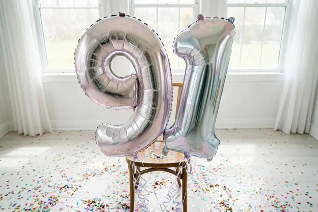 Twee grote zilveren folieballonnen vormen 91 aan een houten stoel in een lichte kamer met witte muren, ramen en gekleurde confetti op de vloer.