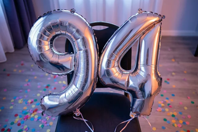 Zilveren folieballonnen vormen 94 aan een zwarte stoel in een lichte kamer met gordijnen en kleurrijke confetti op de houten vloer.