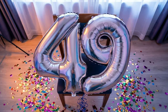 Zilveren folieballonnen 4 en 9 zijn vastgemaakt aan een stoel in een lichte kamer met kleurrijke confetti op de houten vloer en pastelkleurige gordijnen.