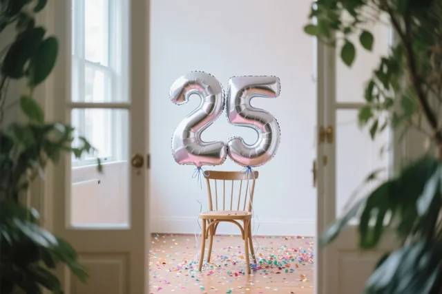 Twee zilveren folieballonnen vormen 25 aan een houten stoel in een lichte kamer met confetti op de vloer en groene planten aan weerszijden.