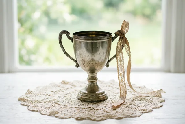 Een oude zilveren trofee met een lint staat op een kanten kleedje op een wit houten oppervlak, met een helder raam op de achtergrond.