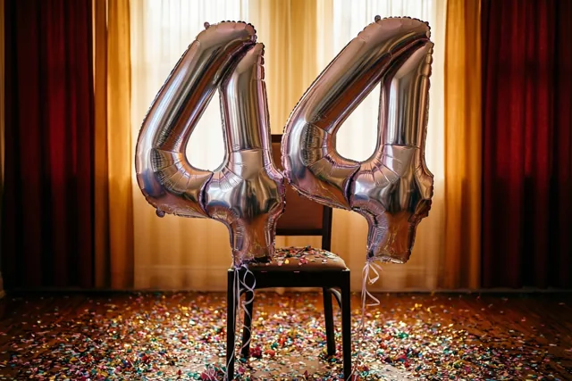 Twee grote zilveren folieballonnen vormen 44 aan een stoel, met confetti op de houten vloer en rode gordijnen bij een licht raam op de achtergrond.