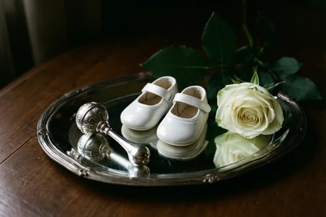 Een spiegelend zilveren dienblad met witte babyschoentjes, een zilveren rammelaar en een witte roos met groene bladeren staat op een donkere houten tafel.