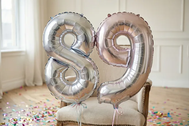 Zilveren en roségouden folieballonnen in de vorm van 89 hangen aan een stoel in een lichte kamer met confetti op de houten vloer.