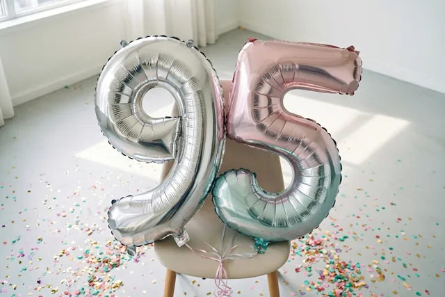 Twee grote zilveren en roségouden folieballonnen in de vorm van 9 en 5 zijn aan een stoel vastgemaakt in een lichte, sober ingerichte kamer met kleurrijke confetti op de vloer.