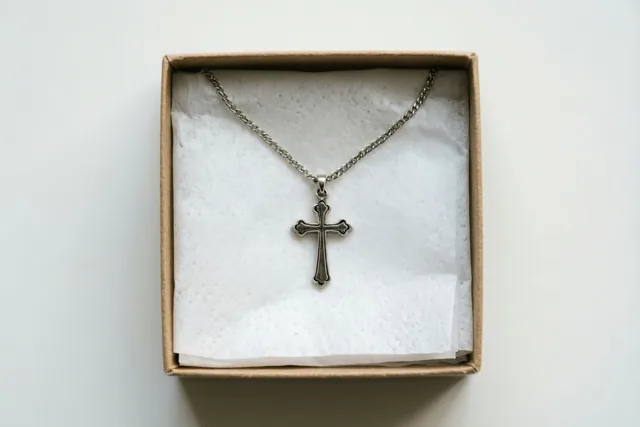 Een zilveren halsketting met een kruis ligt op wit zijdepapier in een open kartonnen geschenkdoos tegen een witte achtergrond.