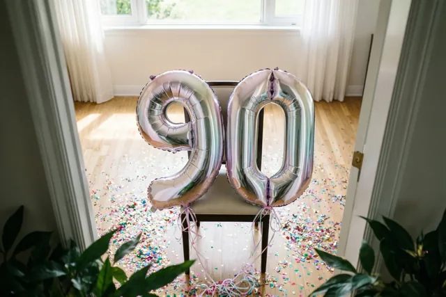 Zilveren en pastelkleurige cijferballonnen vormen 90 aan een stoel in een lichte kamer met confetti op de houten vloer en daglicht door een raam op de achtergrond.