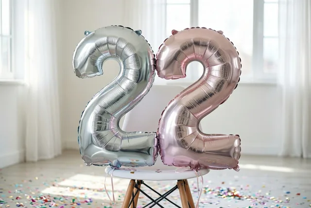 Een zilveren en een roségouden cijferballon vormen 22 aan een witte stoel in een lichte kamer met confetti op de vloer en witte gordijnen.