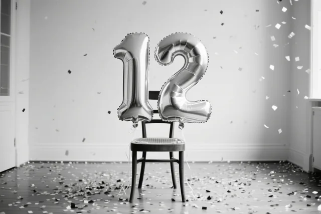 Twee grote zilverkleurige folieballonnen vormen 12 aan een houten stoel in een lichte, sober ingerichte kamer, met confetti op de vloer en in de lucht.