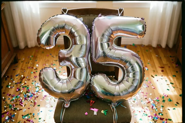 Twee grote zilveren folieballonnen in de vorm van 3 en 5 zijn vastgemaakt aan een stoel in een lichte kamer met kleurrijke confetti op de houten vloer.