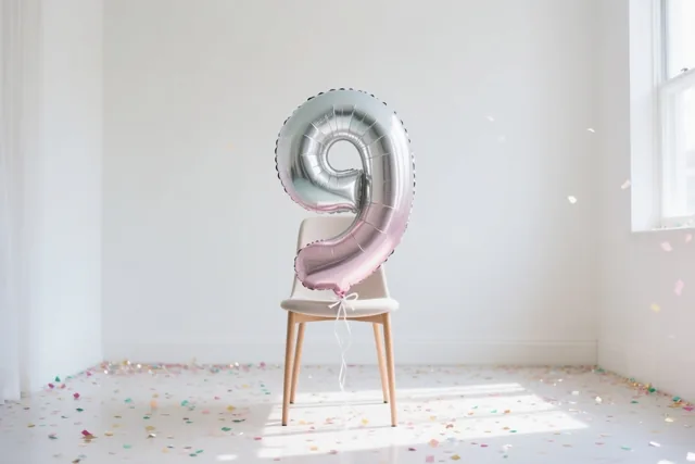 Een grote zilveren en pastelroze ballon in de vorm van een 9 is vastgemaakt aan een lichtgekleurde stoel in een lichte, sobere kamer, met kleurrijke confetti op de vloer en daglicht vanuit een raam rechts.