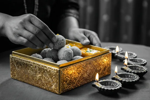 Handen leggen ronde ladoos en vierkante barfi in een sierlijke gouden geschenkdoos op een donkere tafel, verlicht door een rij brandende olielampjes.