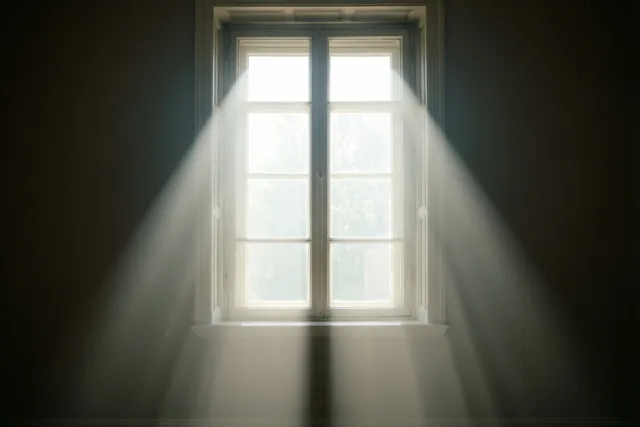 Felle zonnestralen vallen neer vanuit een hoog raam met witte kozijnen in een donkere muur.
